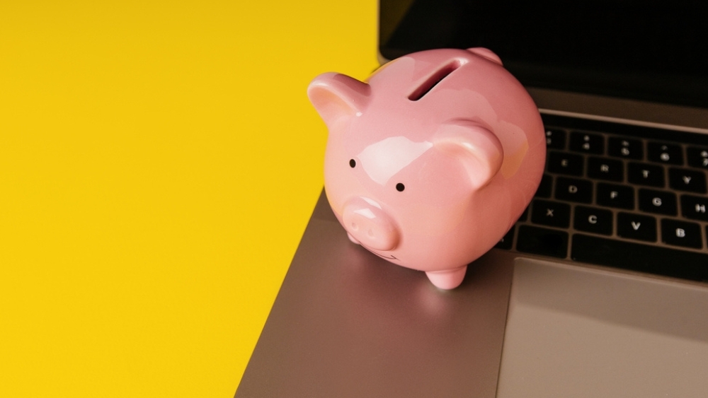 Pink piggy bank perched on a laptop corner, set against a yellow background