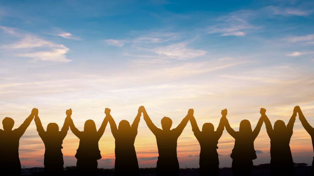 Silhoettes of lots of people holding raised hands against the sunset, representing community
